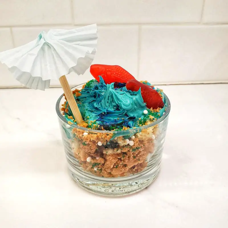 clear glass filled with graham cracker crumbs, blue icing, swedish fish, and a white umbrella