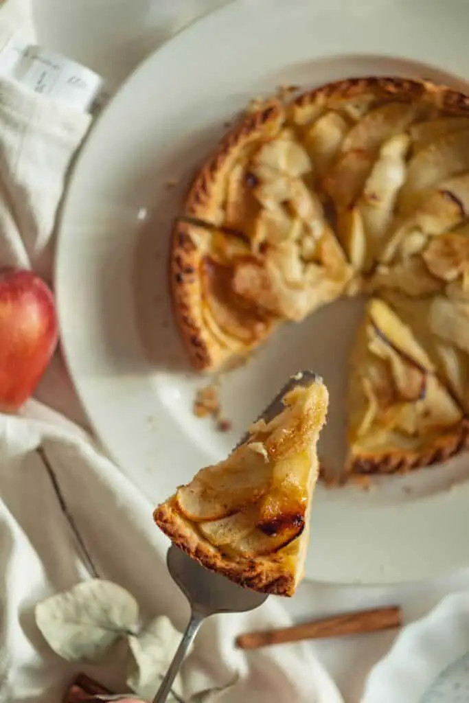 one slice of apple pie raised above an apple pie on a plate with a towel and apple beside it